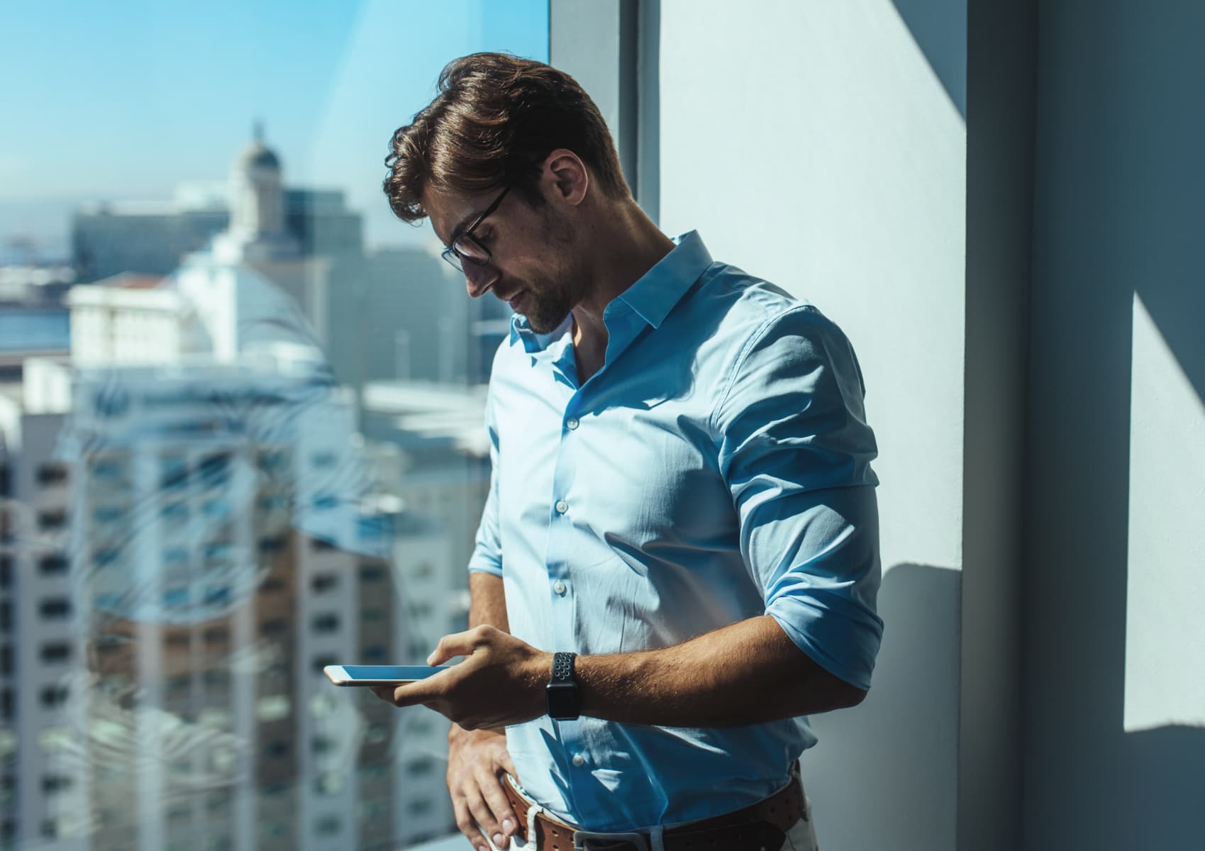 a man standing in front of a window in a city, looking at a smarphone, illustrating an article about mobile app cost