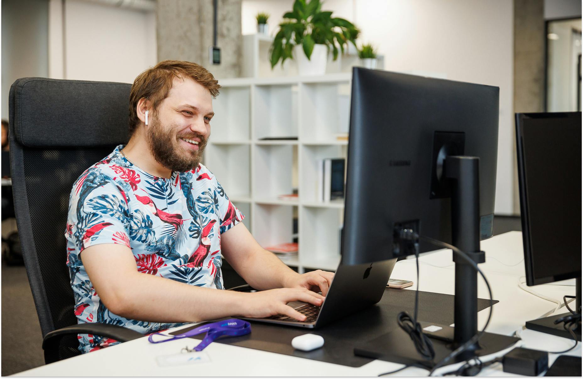Obraz przedstawiający uśmiechniętego programistę siedzącego przed monitorem komputera, piszącym na klawiaturze lapopta